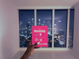 Person holding a pink book titled 'The Marketing Burn Book' in front of a city skyline at night.