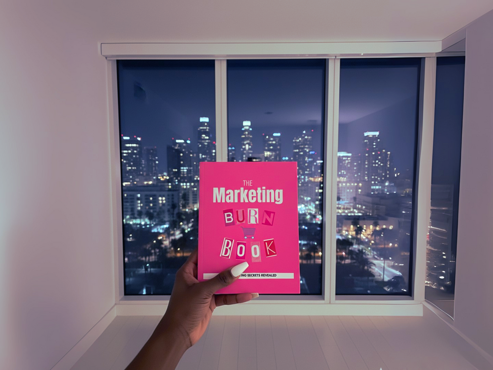 Person holding a pink book titled 'The Marketing Burn Book' in front of a city skyline at night.